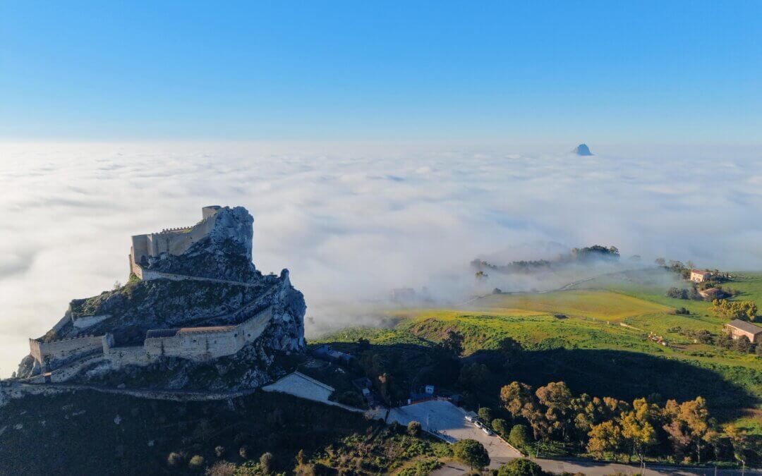 Rocky hillside fortress ruin atop a cliff above a sea of clouds, with a clear blue sky and a distant island on the horizon.