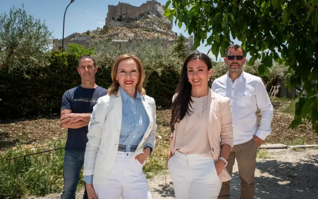 Four smiling adults pose outdoors in a sunny garden with a stone castle ruin on a hill behind them.
